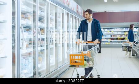 Im Supermarkt: Handsome man schiebt Shopping Card und stösst in der Frozen Goods Section nach Produkten. Der Mann schaut in den Kühlschrank mit Glastür und schaut Stockfoto