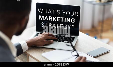 Unkenntlich schwarzen Geschäftsmann arbeiten auf Laptop mit Worten, WAS IST IHR MARKETINGSTRATEGIE auf dem Bildschirm Stockfoto