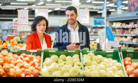 Im Supermarkt: Das glückliche junge Paar wählt Bio-Obst in der Frischwarenabteilung des Stores. Ehemann Benutzt Smartphone, Frau Holt Obst. Stockfoto