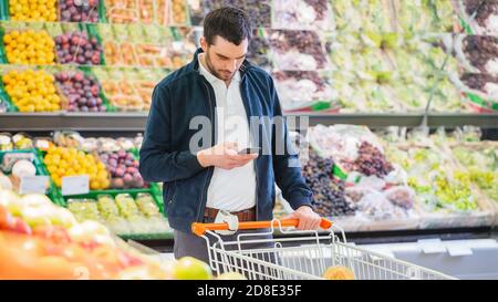 Im Supermarkt: Handsome man mit Smartphone, schiebt den Warenkorb, geht vorbei an Frischwaren Sektion des Stores. Mann im Internet Stockfoto