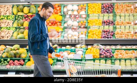 Im Supermarkt: Handsome man mit Smartphone, schiebt den Warenkorb, geht vorbei an Frischwaren Sektion des Stores. Mann im Internet Stockfoto