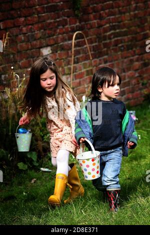 Zwei Kinder mit Eimern auf der Suche nach Ostereiern im Garten. Zu Hause in Wales. An einem warmen Märztag jagen zwei kleine Kinder auf einer Ostereierjagd auf dem Stockfoto