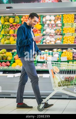 Im Supermarkt: Handsome man mit Smartphone, schiebt den Warenkorb, geht vorbei an Frischwaren Sektion des Stores. Mann im Internet Stockfoto