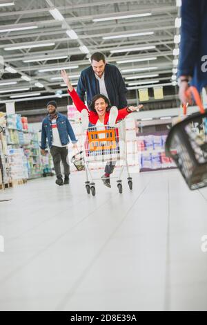 Im Supermarkt: Der Mann schiebt den Einkaufswagen mit der Frau, die darin sitzt. Happy Couple hat Spaß beim Rennen auf einem Trolley durch die frischen Produkte Stockfoto