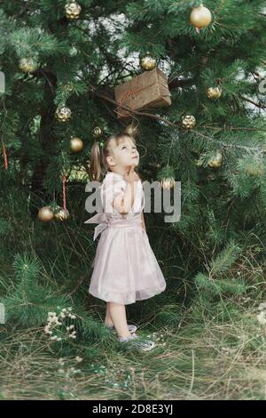 Ein süßes kleines Mädchen in einem rosa Kleid steht am Weihnachtsbaum und versucht zu erraten, was in der Geschenkbox ist. goldenes Geschenk in den Zweigen der Christm Stockfoto