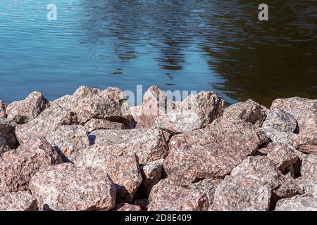 Haufen von großen Steinen aus rosa Granit am Ufer gegen eine blaue Wasseroberfläche. Stockfoto