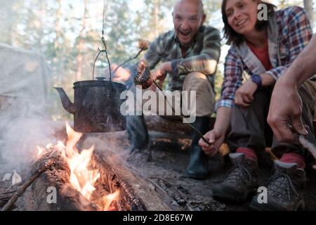 Die Gesellschaft der zufriedenen Freunde braten süsse Marshmallows über dem Feuer mit dem Kessel, der über der Flamme hängt, in einem Campinglager Stockfoto