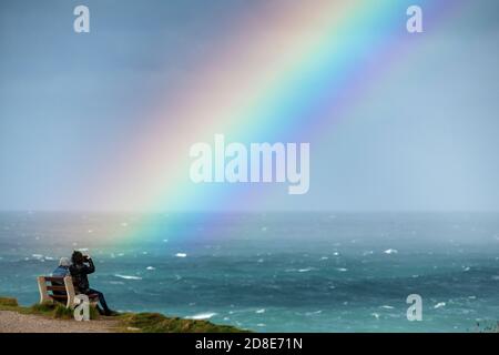 Ein Mann fotografiert mit seinem Smartphone einen Regenbogen über dem Meer, während er auf einer Bank sitzt. Stockfoto