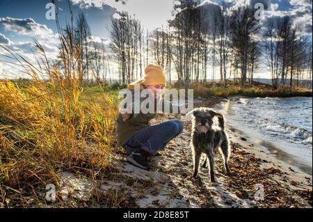 Junge, fröhliche und lächelnde Frau, die im Herbst mit ihrem flauschigen bärtigen Hund am See spielt. Wandern mit Haustieren, freudige Kommunikation mit dem Hund Stockfoto