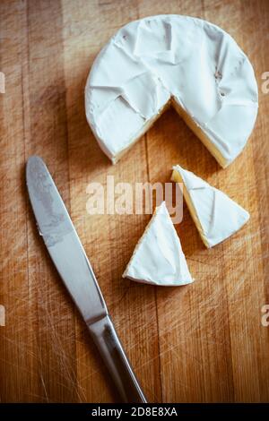 Camembert Käse mit einer dreieckigen Scheiben und einem schmutzigen Messer auf einem Holzbrett. Stockfoto