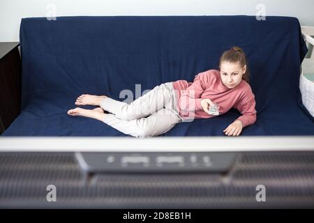 Mädchen im Jugendalter, das Fernsehen schaut, auf der Couch mit tv-Fernbedienung liegend, Breitbild im Vordergrund Stockfoto