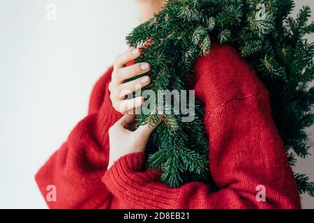 Auf der Schulter einer Frau hängt ein natürlicher Fichtenkranz in einem roten Pullover. Weihnachten minimal Feier Konzept. Stockfoto