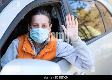 Porträt einer erwachsenen Frau ein Fahrer, der in ihrem Auto sitzt, mit der Hand winkt, Person, die medizinische Maske trägt, um eine Coronavirus-Infektion zu verhindern Stockfoto