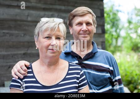 Porträt des älteren Paares mit Ehemann liegt Hand auf Frau Schulter, lächelnd kaukasischen älteren Menschen im Freien neben Holzhaus stehen Stockfoto
