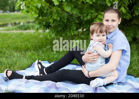 Portrait eines jungen Mädchens, das ihre kleine Schwester umarmt, Mädchen, die auf einer Decke auf grünem Gras in einem Park sitzen Stockfoto