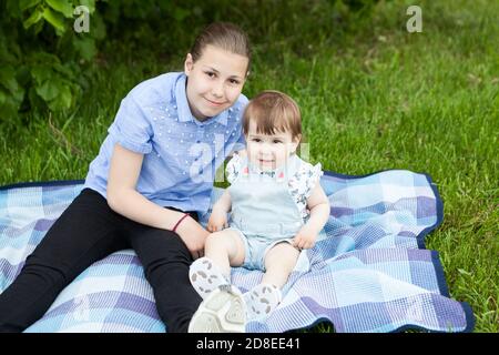 Zwei kaukasische Schwestern unterschiedlichen Alters sitzen zusammen auf Decke in grünem Gras, glücklich und lächelnd Blick auf die Kamera Stockfoto