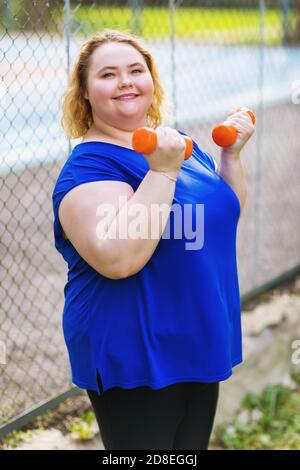 Portrait einer positiven plus size blonde macht Übungen mit Hanteln auf dem Hintergrund des Sportplatz. Gesundes Lifestyle-Konzept Stockfoto