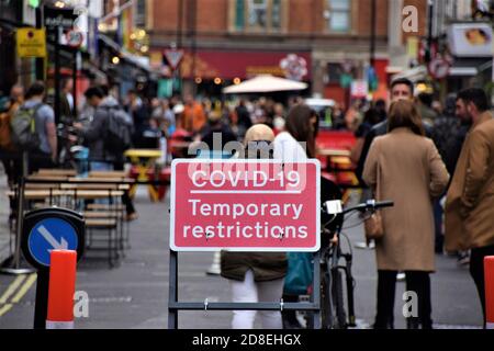 London, Großbritannien. Oktober 2020. Covid-19 temporäre Einschränkungen Straßenschild in Soho, mit einer verschwommenen kleinen Menschenmenge im Hintergrund.Neue Sperrstunde Beschränkungen in der Hauptstadt haben dazu geführt, dass weniger Kunden Restaurants und Bars besuchen. Kredit: Vuk Valcic/SOPA Images/ZUMA Wire/Alamy Live Nachrichten Stockfoto