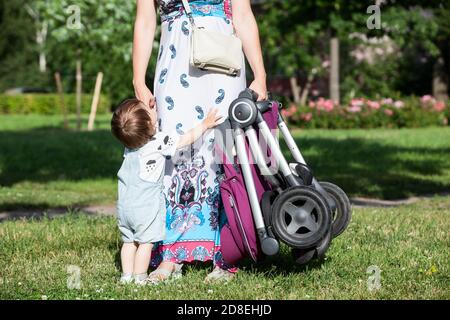 Launisch Kind bittet seine Mutter, in den Armen zu nehmen, unkenntlich Frau hält Baby Kinderwagen in der Hand, Sommerwiese im sonnigen Park Stockfoto