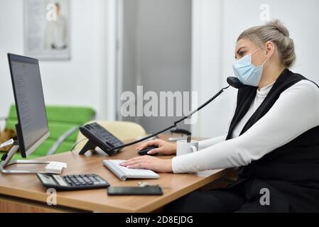 Junge Verkaufsleiterin in einer Gesichtsmaske im Gespräch Das Telefon im Büro Stockfoto