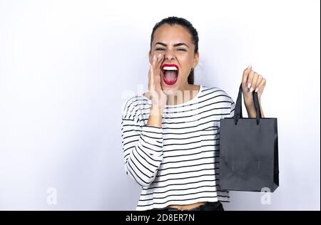 Junge schöne Frau hält eine schwarze Einkaufstasche und schreit laut zur Seite mit den Händen am Mund. Schwarzer Freitag Stockfoto