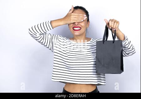 Junge schöne Frau mit einer schwarzen Einkaufstasche Junge schöne Frau hält Geschenke über isolierten weißen Hintergrund ernst und Abdeckung Ihre Augen wi Stockfoto