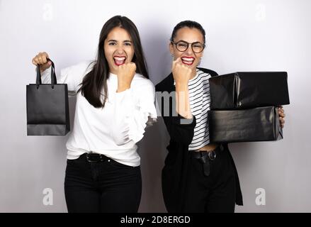 Zwei junge schöne Frauen mit schwarzen Einkaufstaschen und Boxen aufgeregt machen Gewinnergeste mit erhobenen Armen, lächelnd und schreien nach Erfolg. Bla Stockfoto