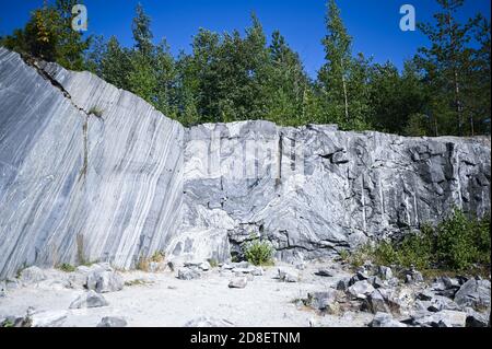 Felsenschnitt rohe Oberfläche aus weißem und grauem Marmor in der natürlichen Umgebung mit Bäumen und blauen Himmel. Blaue Marmor Karriere Textur. Hintergrund Stein, industrielles Material für Gebäude und Interieur. Stockfoto