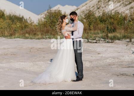 Junge liebende Paar in medizinischen Masken im Park während der Quarantäne an ihrem Hochzeitstag zu Fuß. Stockfoto