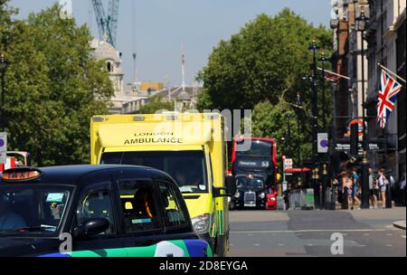 Stadtperspektive entlang einer belebten Straße im Zentrum von London mit Taxi, gelber Ambulanz und rotem Bus im Hintergrund. Englische Flagge an einem Gebäude Stockfoto