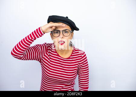 Junge schöne Brünette Frau trägt französisch Baskenmütze und Brille auf weißem Hintergrund sehr glücklich und lächelnd Blick weit weg mit Hand über Kopf. Meer Stockfoto