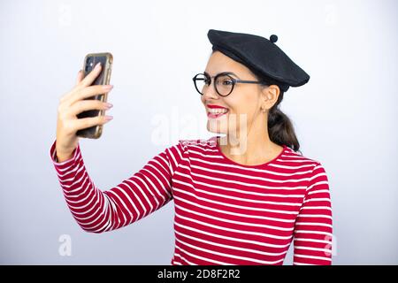 Junge schöne Brünette Frau trägt französisch Baskenmütze und Brille über Weißer Hintergrund nimmt ein Selfie mit ihrem Handy Stockfoto