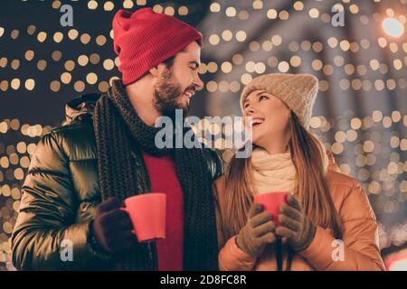 Foto von liebevollen Paar halten heißen Kaffee Getränk Tasse unter Weihnachten Abendbeleuchtung tragen Saison Tradition Mäntel Stockfoto