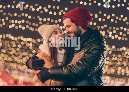 Foto von Liebespaar Umarmung unter Weihnachten Abend im Freien Lichter Tragen Saison Winter Mäntel Kopfbedeckung Stockfoto