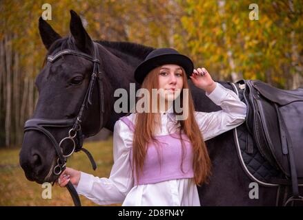 Rothaarige Mädchen in einem schwarzen Hut mit einem Pferd im Herbstwald. Stockfoto