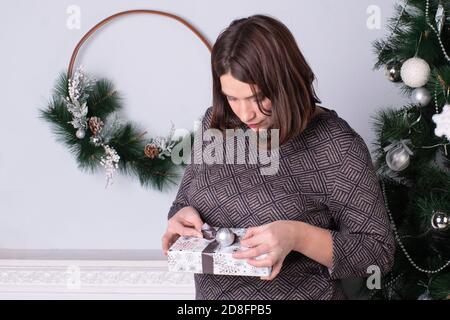 Schöne glückliche Frau sitzt neben einem Weihnachtsbaum mit einem Geschenk in den Händen, eine Frau öffnet ein Neujahrsgeschenk Stockfoto