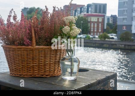 Korbkorb mit Wildblumen und transparenter Vase auf einem Holztisch am Fluss in der Stadt. Schöne Einrichtung. Nahaufnahme, selektiver Fokus, Unschärfe. Stockfoto