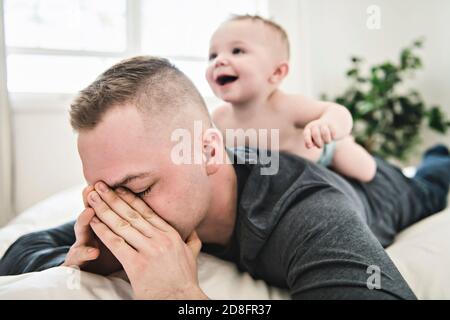 Müde Vater schlafen, aber sein Baby Junge stört ihn. Junger glücklicher Vater liegt auf dem Bett, während sein Sohn mit ihm spielt Stockfoto