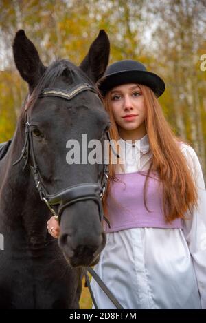 Rothaarige Mädchen in einem schwarzen Hut mit einem Pferd im Herbstwald. Stockfoto