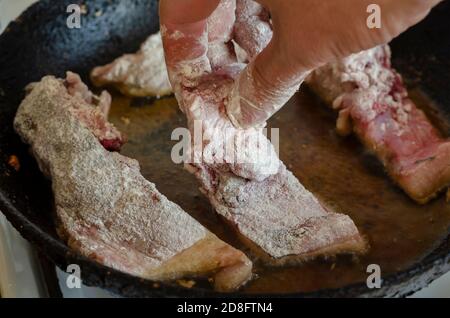 Männliche Hand legt Stücke von rohem Fisch in Mehl gerollt in der heißen Pfanne. Fisch in gusseiserner Pfanne anbraten. Kochen von hausgemachten Mahlzeiten. Stockfoto
