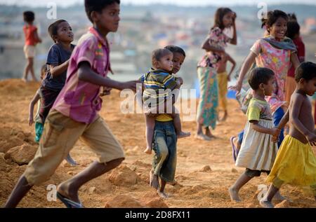 Rohingya-Menschen gehen am 16. April 2018 im Flüchtlingslager Kutupalong in Maynar Guna, in der Nähe von Cox's Bazar, Bangladesch, herum, als Unterstände hinter ihnen zu sehen sind. Rohingya Menschen, die vor der Unterdrückung in Myanmar flohen, versuchen, in Notsiedlungen aus Bambus, adobe oder Nylon im Flüchtlingslager Kutupalong unter harten Bedingungen zu leben. Mehr als 650,000 Rohingya haben seit August letzten Jahres die Grenze zu Bangladesch überschritten und sind vor der Gewalt geflohen. Stockfoto