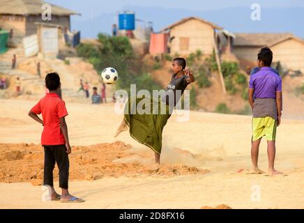 Rohingya spielt Fußball, als am 16. April 2018 im Flüchtlingslager Kutupalong in Maynar Guna in der Nähe von Cox's Bazar in Bangladesch Unterkünfte hinter ihnen zu sehen sind. Rohingya Menschen, die vor der Unterdrückung in Myanmar flohen, versuchen, in Notsiedlungen aus Bambus, adobe oder Nylon im Flüchtlingslager Kutupalong unter harten Bedingungen zu leben. Mehr als 650,000 Rohingya haben seit August letzten Jahres die Grenze zu Bangladesch überschritten und sind vor der Gewalt geflohen. Stockfoto