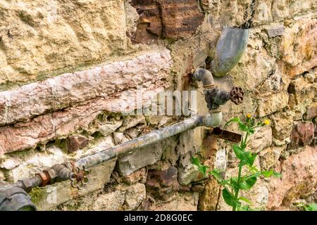 Alte rostige Wasserventil mit Rohren auf dem Hintergrund Eine zerstörte Steinmauer eines verlassenen Hauses Stockfoto