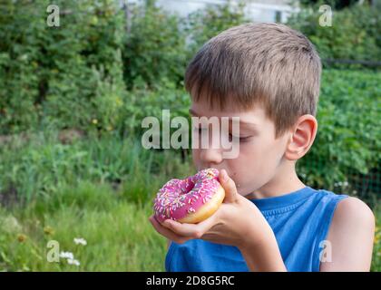 Kaukasischer Junge isst rosa Donut. Ein Kind hält einen süßen Leckerbissen im Park. Ungesunde Ernährung Konzept, Naschen süße Lebensmittel Stockfoto