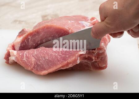 Nahaufnahme Hand hält Messer und schneidet rohes Schweinefleisch auf Ein schneidendes Whiteboard auf dem Küchentisch Stockfoto