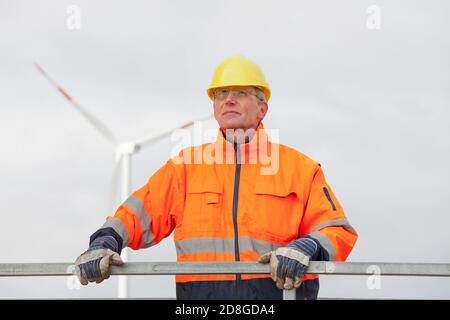 Lächelnder reifer Ingenieur mit Hut und Schutzkleidung in Vorderseite der verschwommenen Windenergieanlage im Hintergrund Stockfoto