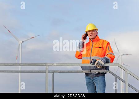 Techniker mit Schutzarbeiten tragen Gespräch auf Mobiltelefon in Vorderseite der Windenergieanlagen im Hintergrund Stockfoto