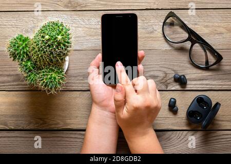 Smartphone mit schwarzem Bildschirm Display in weiblichen Händen, kabellose Kopfhörer, Kaktus, Ladegerät Kapsel, Tasse Kaffee auf Holztisch Flat Lay Draufsicht Stockfoto