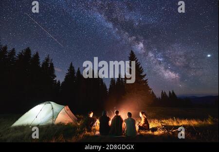 Abend Sommer Camping, Fichtenwald im Hintergrund, Himmel mit fallenden Sternen und Milchstraße. Gruppe von fünf Freunden, die zusammen am Lagerfeuer in den Bergen sitzen und die frische Luft in der Nähe des beleuchteten Zeltes genießen Stockfoto