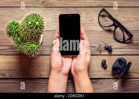 Smartphone mit schwarzem Bildschirm Display in weiblichen Händen, kabellose Kopfhörer, Kaktus, Ladegerät Kapsel, Tasse Kaffee auf Holztisch Flat Lay Draufsicht Stockfoto